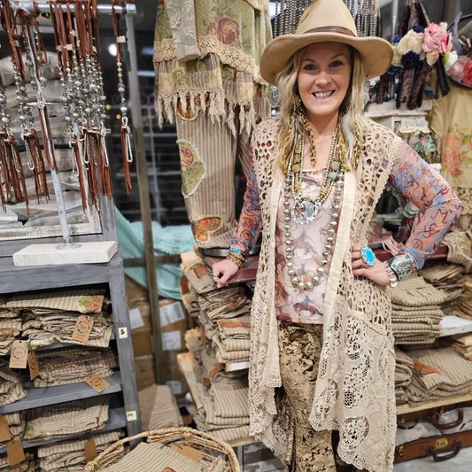 Woman in a store wearing a hat and a crocheted sleevless cardigan, surrounded by decorative items.