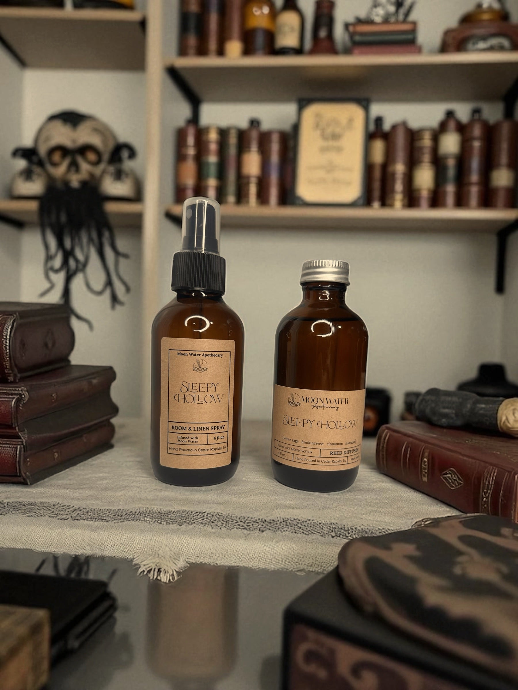 Two brown Sleepy Hollow Room Spray & Reed Diffuser bottles by Moon Water Apothecary sit among vintage books. Behind them are more books, a tentacled skull, and scents of cinnamon, lavender, sage & frankincense for a mysterious vibe.
